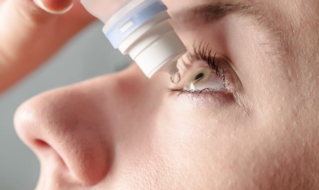 Close up view of young woman applying eye drop to left eye, arti